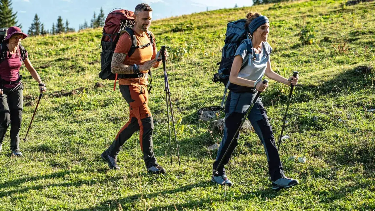 Trzy osoby wędrują po zielonym zboczu. Mężczyzna i kobieta w wygodnych, sportowych spodniach w góry, z plecakami i kijkami trekkingowymi.
