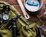 Gotowy **zestaw survivalowy**: plecak, kompas, nóż, latarka, krzesiwo, lina, kuchenka turystyczna, menażka i sztućce.