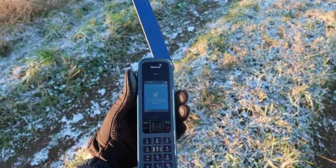 Ręka w rękawiczce trzyma telefon Inmarsat IsatPhone, gotowy do nawiązania połączenia przez telefonię satelitarną w mroźny poranek.