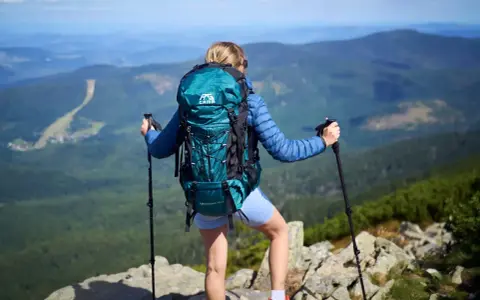 Kobieta z turkusowym plecakiem turystycznym i kijkami trekkingowymi na szczycie góry.
