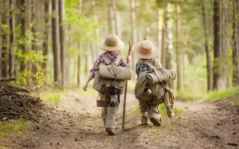 Dwoje dzieci w kapeluszach i z plecakami wyrusza na leśną ścieżkę. To przygoda i nauka survivalu dla dzieci.