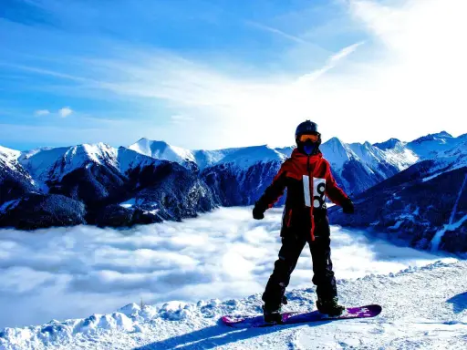 Snowboardzista w kurtce hardshell stoi na szczycie góry, nad morzem chmur. Góry w tle.