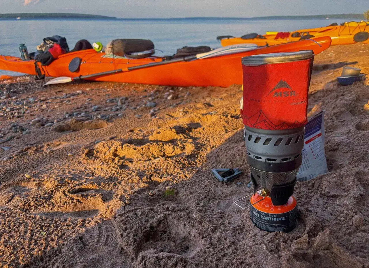 Jaka kuchenka turystyczna MSR na plaży, obok pomarańczowego kajaka. Idealna na wyprawę nad jezioro.
